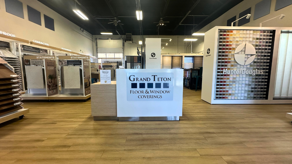 Interior of a Grand Teton Floor & Window Coverings showroom with display racks along the walls, a central counter, and a large sample board on the right showing many carpet, hardwood, and tile swatches.